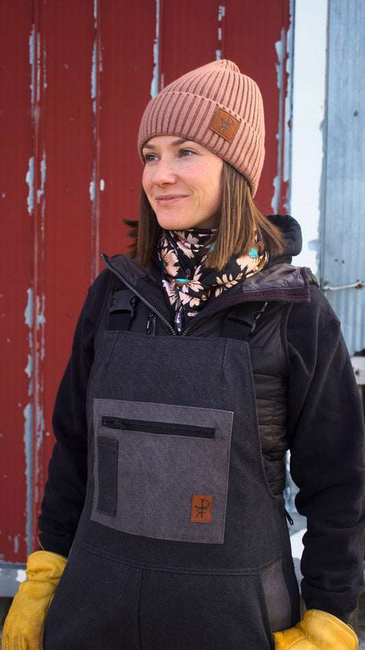 Person wearing a black overalls, pink beanie, and yellow gloves standing in front of a red wall.
