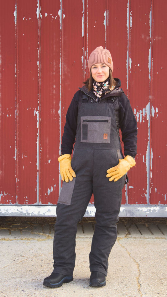 Person wearing a black overall, gloves, and a beanie in front of a red barn.