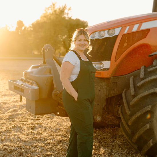 Woman wearing green overalls standing next to a large orange tractor in a field