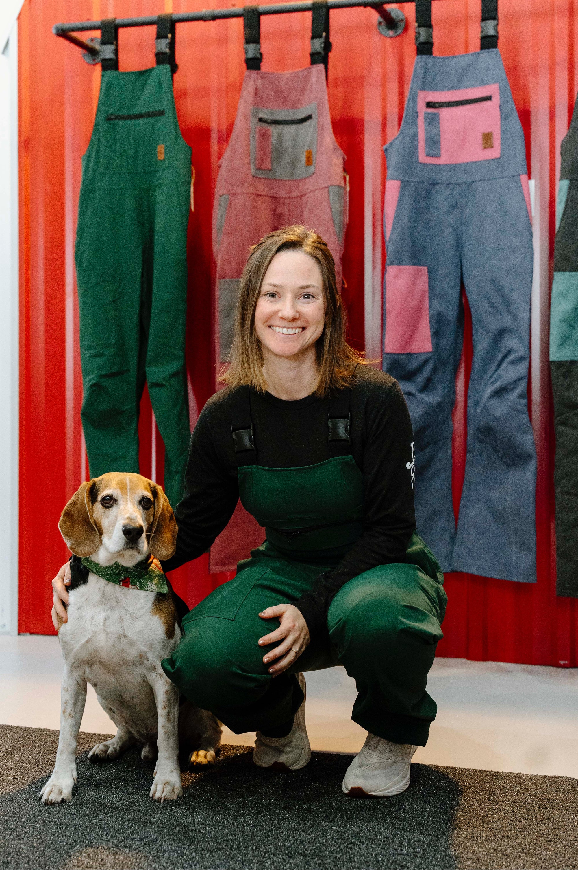 Woman in green overalls kneeling with a dog in front of a display of colorful overalls against a red wall.