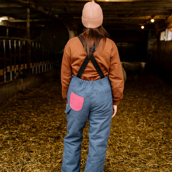 Person wearing a brown shirt, blue overalls, and pink hat standing in a barn.