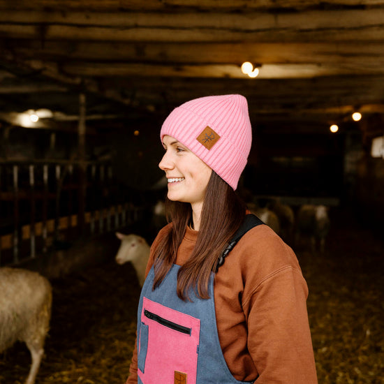 Woman wearing a pink beanie and blue overalls standing in a barn with sheep.