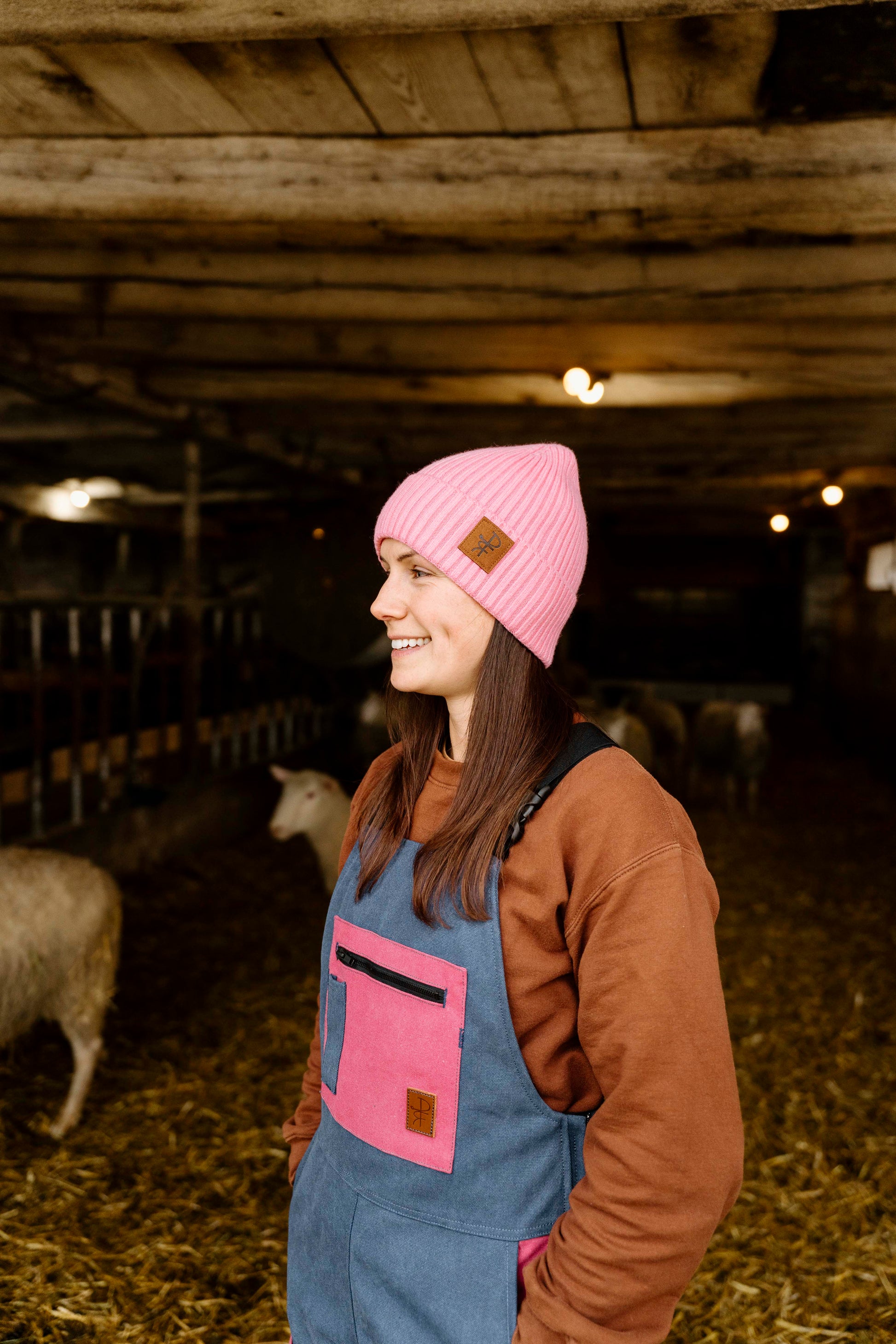 Woman wearing a pink beanie and blue overalls standing in a barn with sheep.
