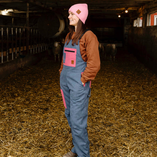 Person wearing a pink beanie and blue overalls standing in a barn with animals in the background.