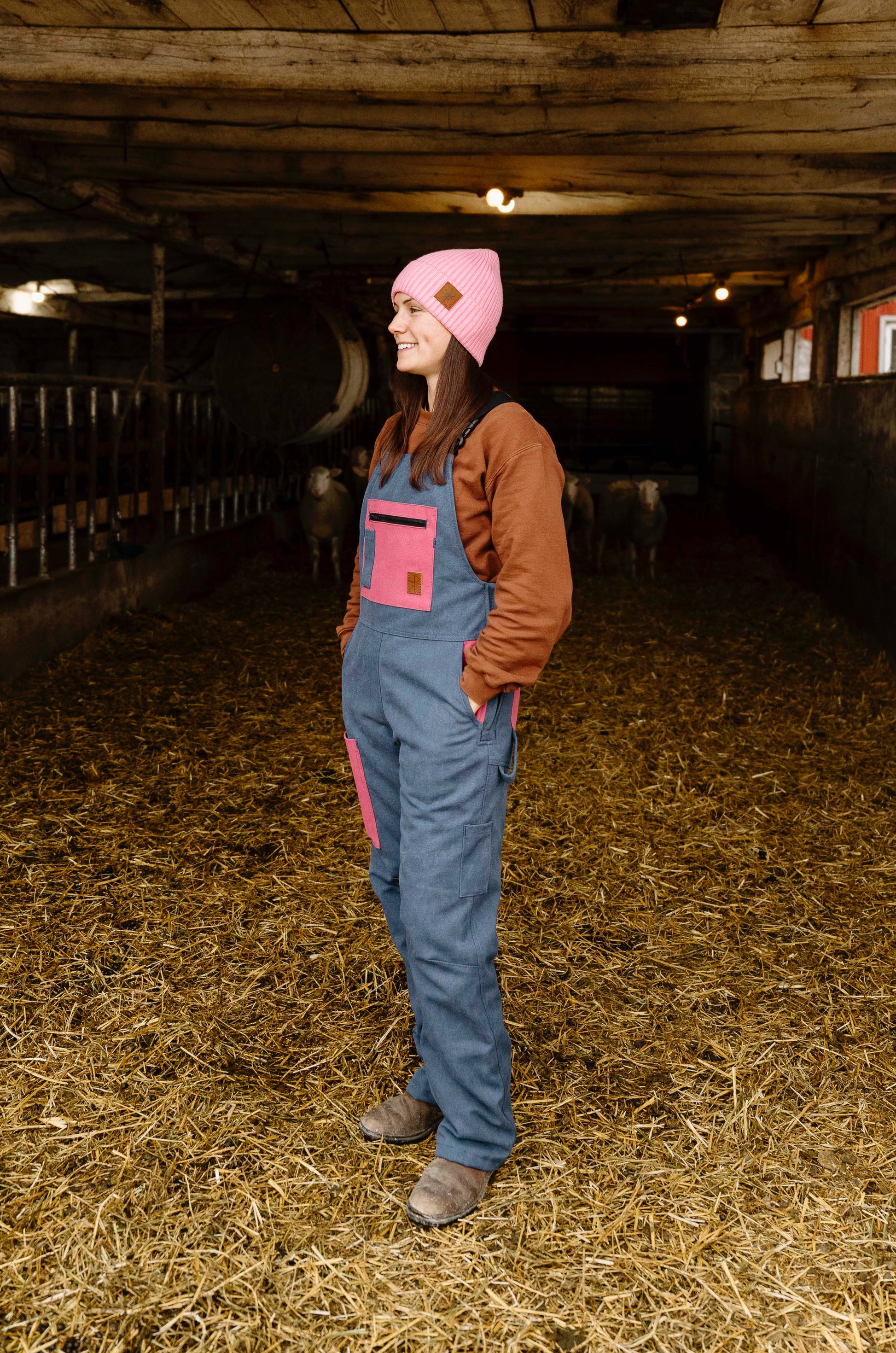 Person wearing a pink beanie and blue overalls standing in a barn with animals in the background.