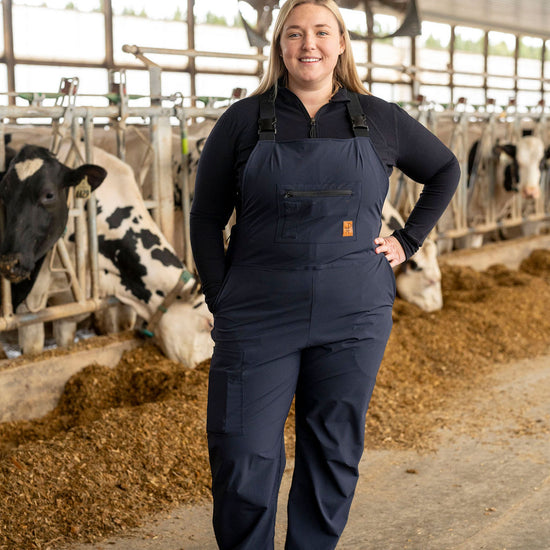 Woman in a blue overall standing in a barn with cows.