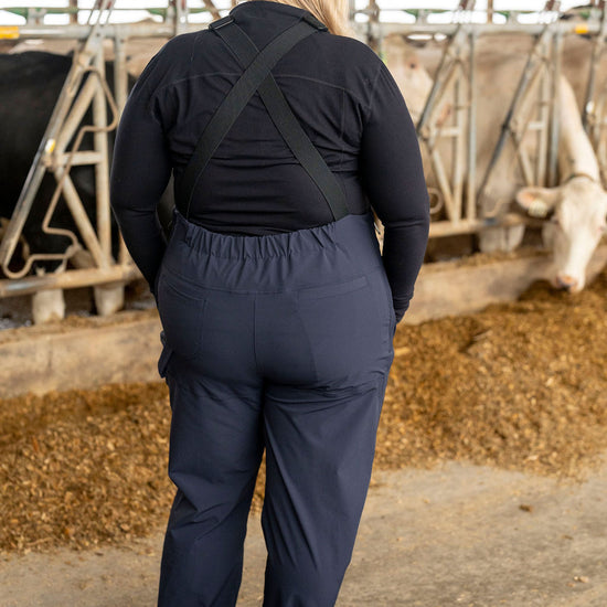Person wearing navy overalls in a barn setting with cows and hay.